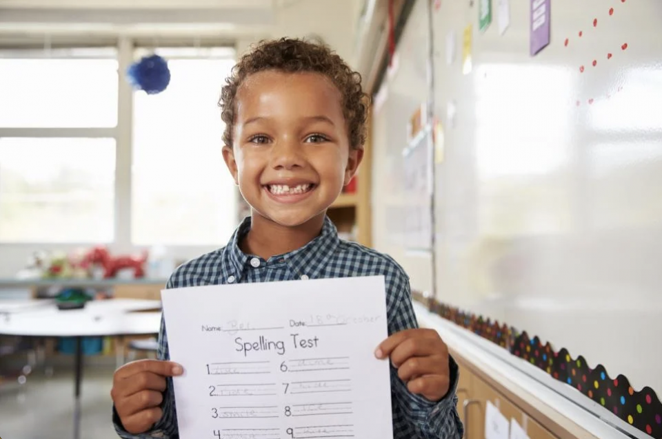 Image of elementary student smiling and holding up a spelling test. 