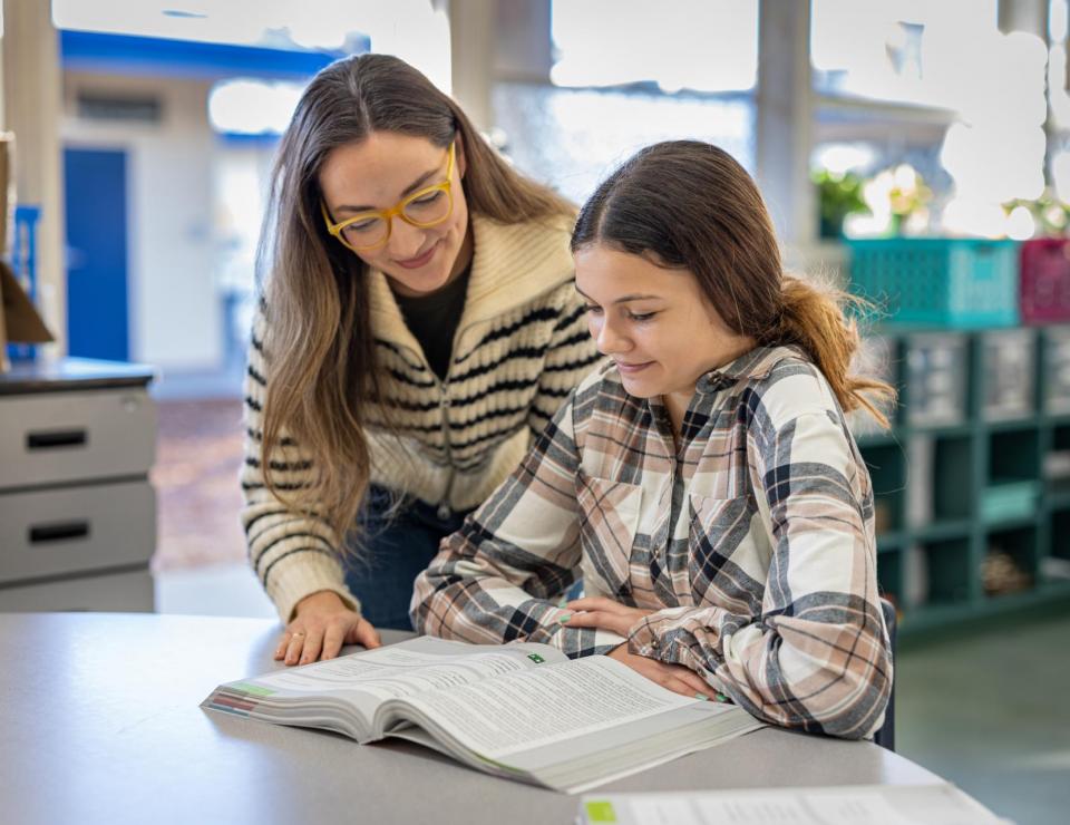 A teacher leans stands over a sitting middle school student in front of an open book. 
